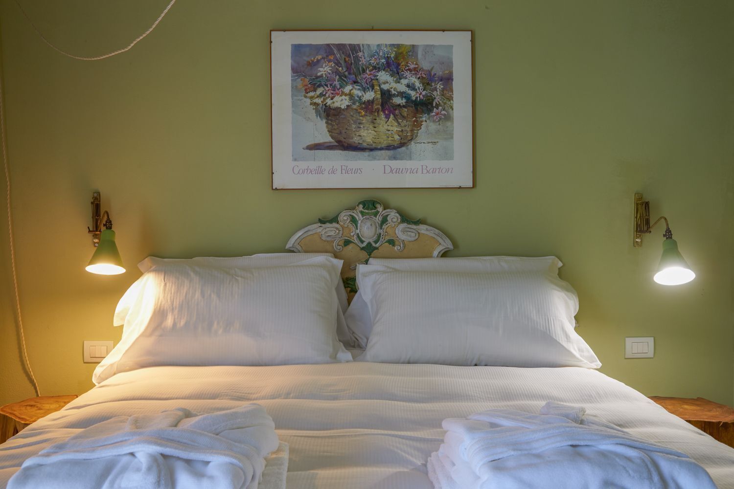 Un letto con lenzuola bianche, due lampade a parete e un quadro raffigurante un cesto di fiori sopra di esso.