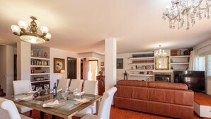 Een eetgedeelte met een glazen tafel, witte stoelen, een kroonluchter, een bruine leren bank en een open haard.