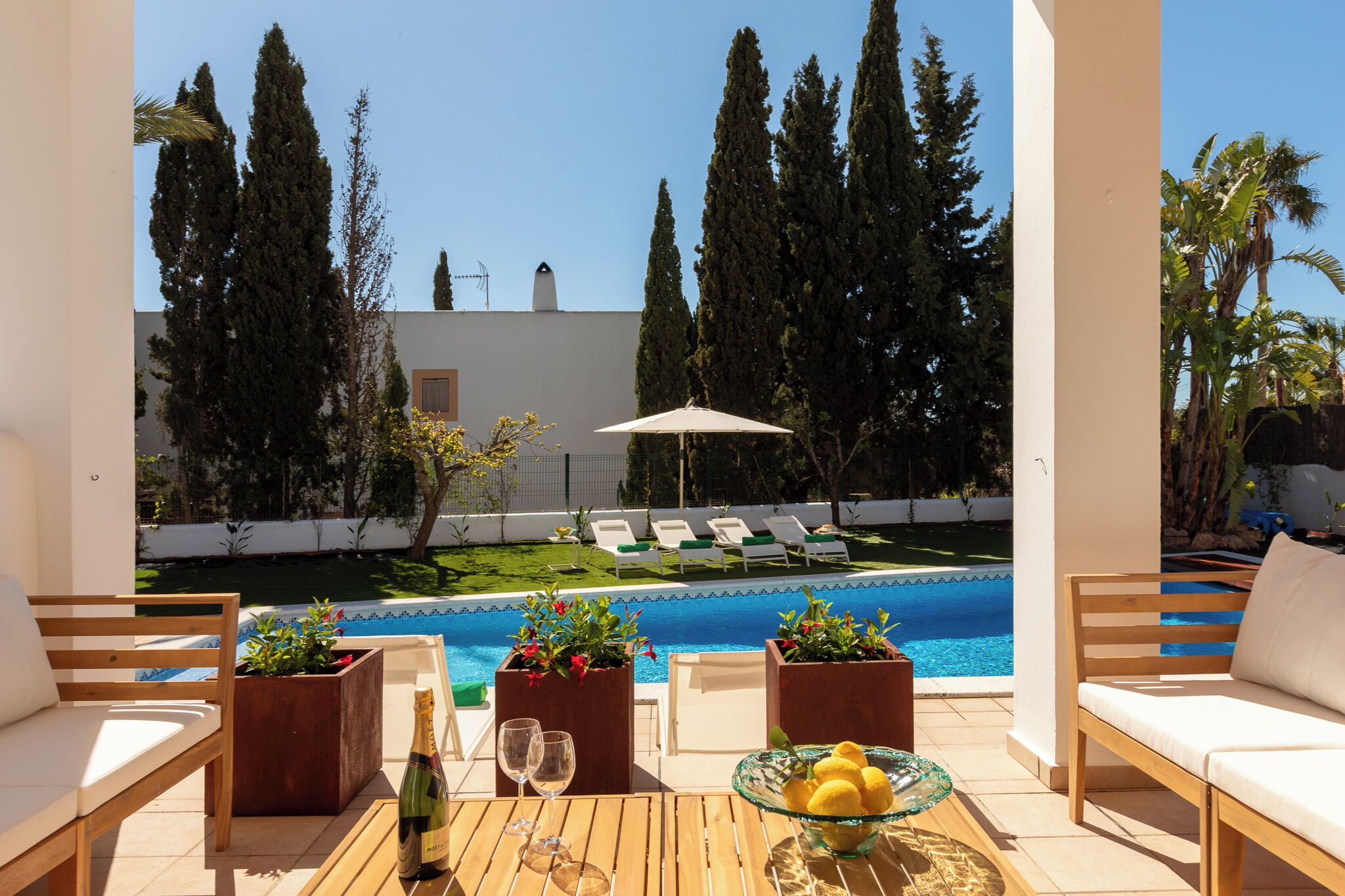A poolside area with lounge chairs, a table with a bottle and glasses, and planters with flowers.