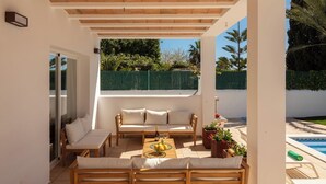 Eine überdachte Terrasse mit einer Couch, Sesseln, einem Tisch mit einer Flasche und einer Schüssel mit Obst.