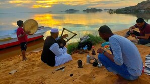 A group of people sitting on a sandy beach with a boat and a calm sea in the background. - Kabara (Abu Simbel)