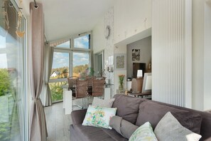 Een woonkamer met een bruine bank, een eethoek met rieten stoelen en uitzicht op het omliggende landschap.