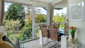 A dining area with wicker chairs, a glass table, and a view of trees through large windows. - Apartment “Wintergarten Am Thingplatz” with Shared Terrace, Garden & Wi-Fi (Rinteln)