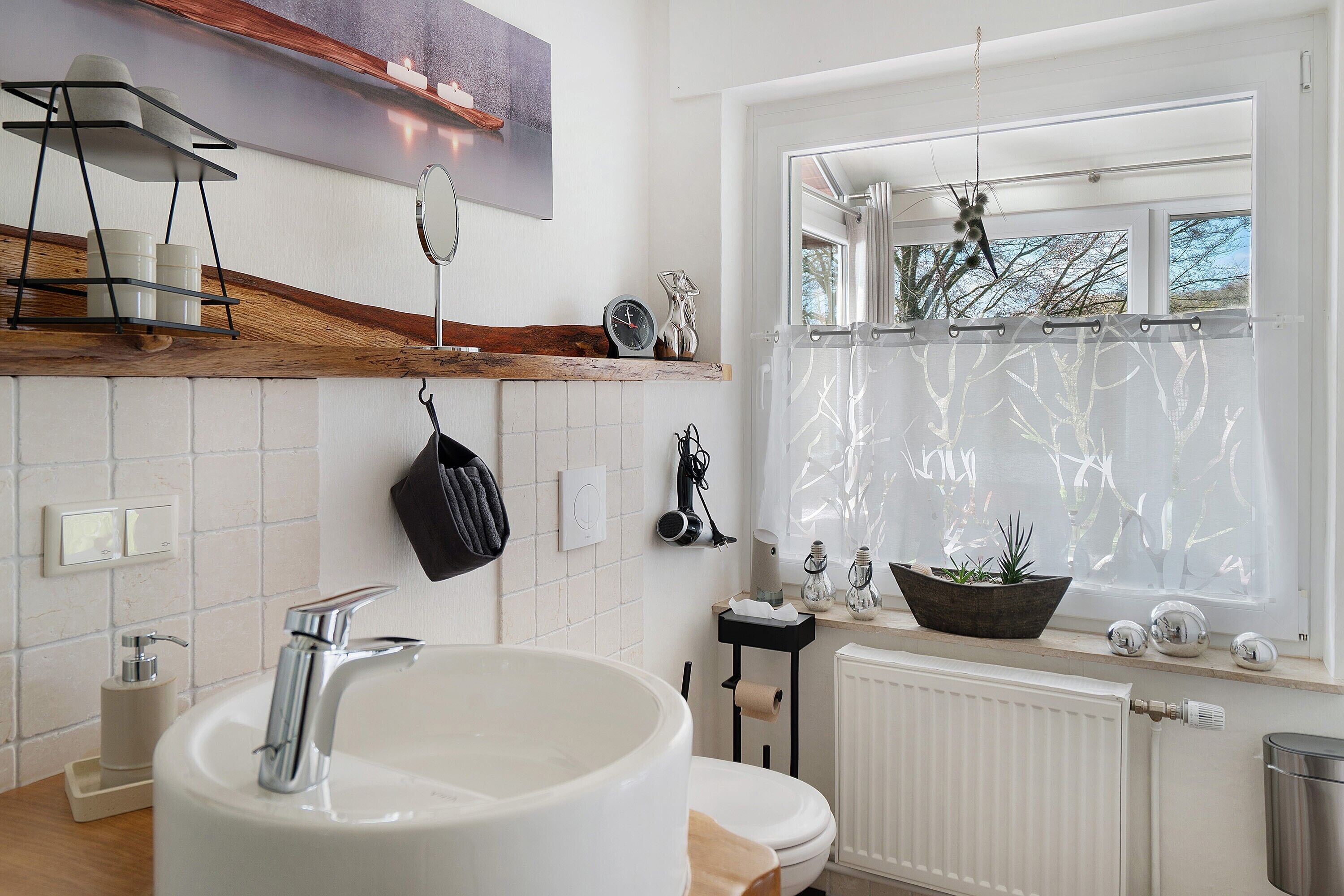 Une salle de bain avec un grand lavabo, un miroir, une étagère contenant des articles de toilette, un radiateur et une fenêtre donnant sur des arbres.