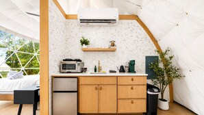 A compact kitchen with a microwave, toaster, and coffee maker, a small refrigerator, wooden cabinets, and a potted plant. - Glamping Collective Chattanooga (Trenton)