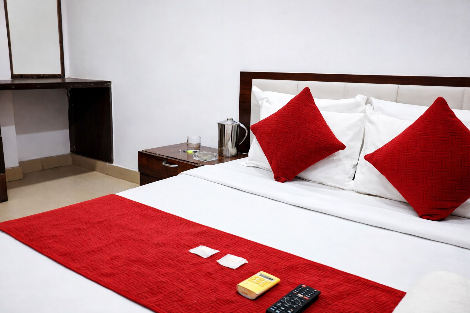 Een hotelkamer met een bed, een nachtkastje, een rood vloerkleed, een afstandsbediening en een kleine fles.