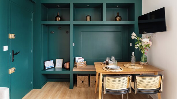 Una habitación color turquesa con una mesa de comedor de madera, sillas de mimbre, un televisor de pantalla plana en la pared y estantes con adornos.
