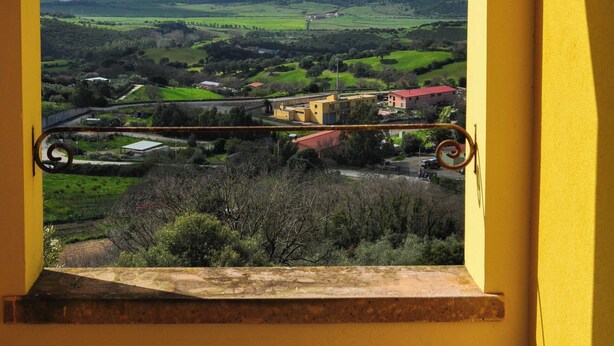 A view through a window with a yellow wall, showcasing a landscape with green fields, a mountain, and scattered buildings. - Il Glicine (Laconi)