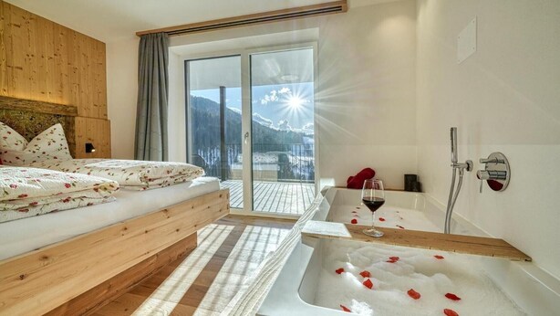Une chambre à coucher avec une tête de lit en bois, une grande fenêtre donnant sur les montagnes, un lit avec une literie à motifs floraux et une baignoire remplie de pétales de roses.
