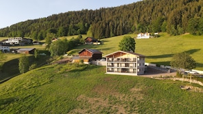 Un immeuble à plusieurs étages doté d’un balcon, entouré de champs verdoyants et d’une colline boisée.