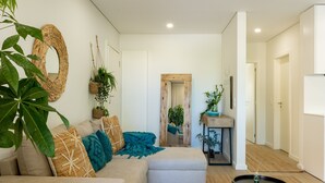 Una sala de estar moderna con un sofá, un reposapiés y una mesa de centro redonda. Hay plantas, un espejo y una puerta que da a otra habitación.