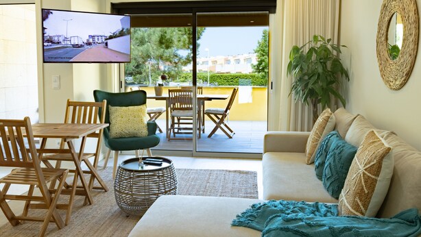 Una sala de estar con un sofá, un televisor montado en la pared, una zona de comedor con mesa y sillas, y un balcón con vistas.