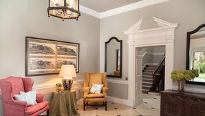 A room with a fireplace, a staircase, a red armchair, a beige armchair, a lamp, and a framed artwork on the wall.