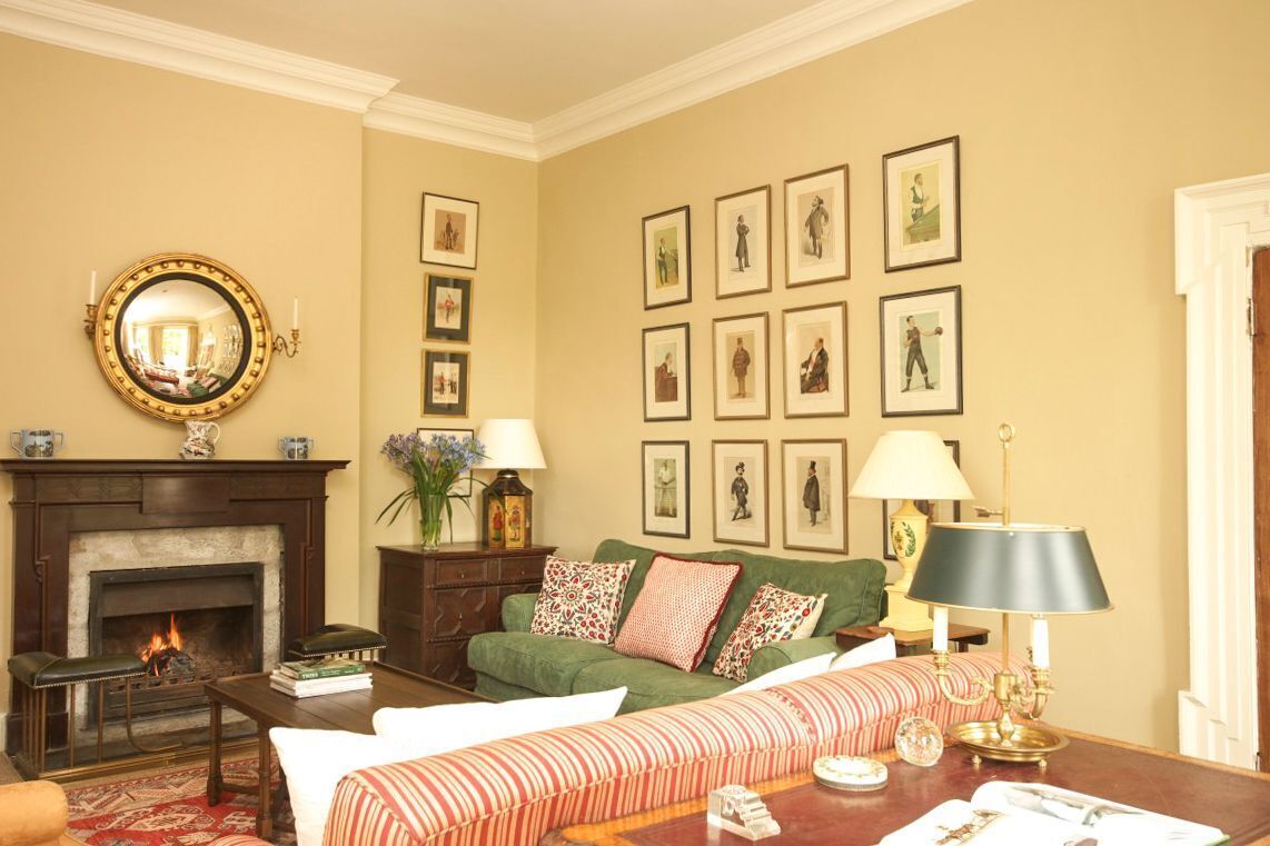 A living room with a fireplace, green sofa, patterned cushions, framed pictures on the wall, and a round mirror above the fireplace.