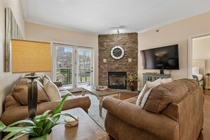A living room with a stone fireplace, a television, and two brown sofas. - 🌲Baskins Creek 206: Cozy Gatlinburg Condo | Balcony & Hot Tub Await🏔️ (Gatlinburg)