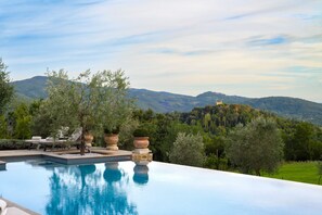 An infinity pool with a view of a castle on a hill, surrounded by olive trees and greenery.
