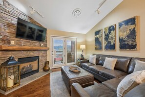 A living room with a stone fireplace, leather sofa, and a flat-screen TV mounted on the wall. - 🏔️Baskins Creek 403 | Cozy Smoky Mountain Retreat | Walk to Downtown🏔️ (Gatlinburg)