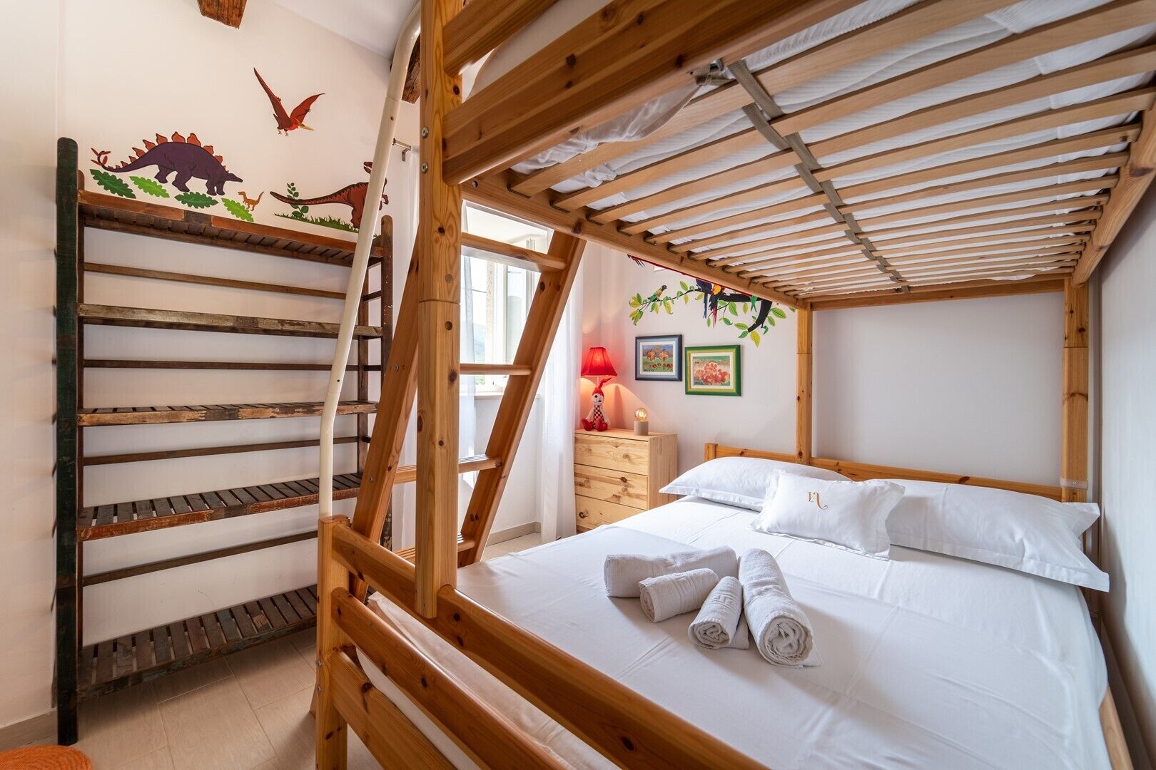 A bunk bed with a ladder, a wooden headboard, a bedside table with a lamp, and a wall with dinosaur decorations.