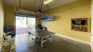 Una habitación con mesa de ping pong, sillas, televisor, una tabla de surf y dardo.