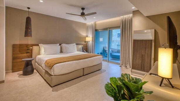 Quarto de hotel moderno com cama, ventilador de teto, janelas amplas e vista para o exterior.