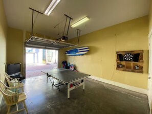 Ein Raum mit Tischtennisplatte, Stühlen, Fernseher, Surfbrett und Dartscheibe.