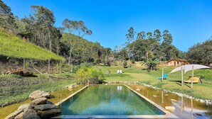 Una piscina rectangular amb aigua cristal·lina, envoltada de roques i vegetació, amb una casa i arbres al fons.