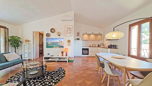 Una sala moderna con sofá, mesa de centro y televisor. Se observa una cocina con comedor y un amplio ventanal.