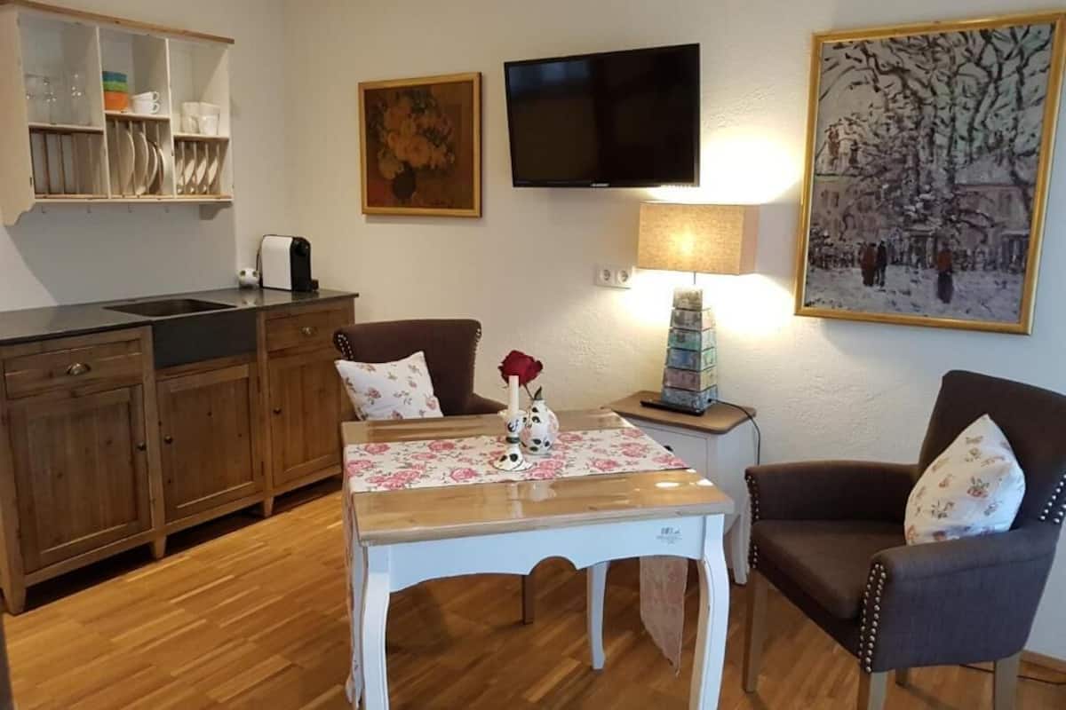 A living room with a dining table, two chairs, a TV, a painting, and a kitchen area with cabinets and a sink.