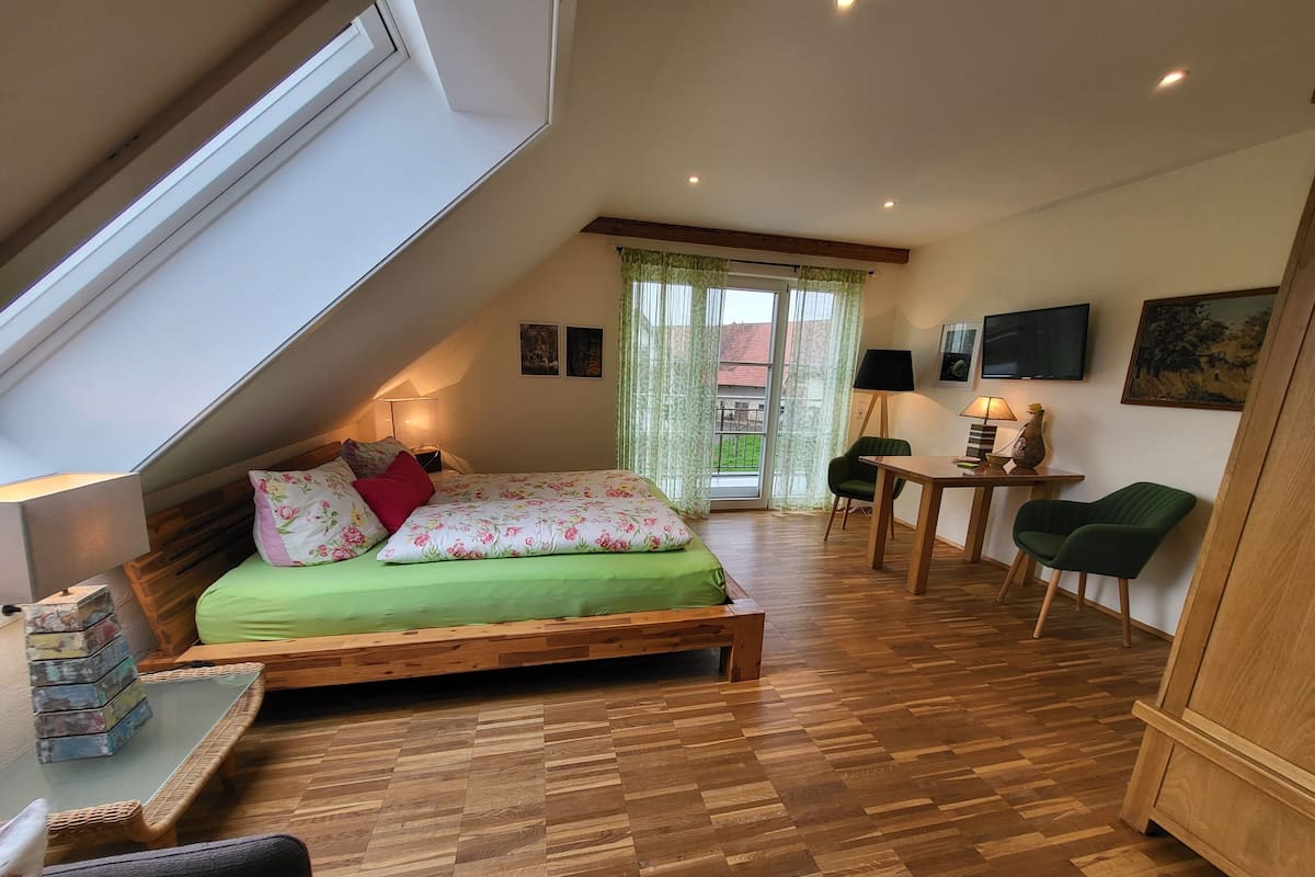 A loft-style bedroom with a sloped ceiling, a bed with floral bedding, a wooden desk, a chair, and a view through a window.