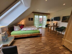 A loft-style bedroom with a sloped ceiling, a bed with floral bedding, a wooden desk, a chair, and a view through a window. - Landgut Apfelrose (Friedrichshafen)