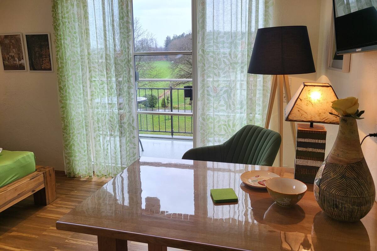 A room with a wooden table, a green chair, a lamp, a vase with a flower, and a view of a balcony with greenery.