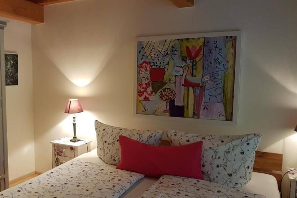 A bedroom with a bed, a nightstand, a lamp, a painting, and a wardrobe.