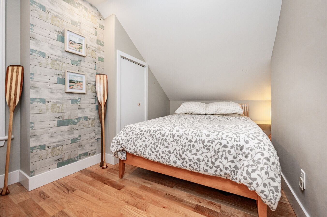 A bedroom with a sloped ceiling, a wooden floor, a bed with a patterned bedspread, a wall with a wooden plank design, and two wooden paddles leaning against the wall.