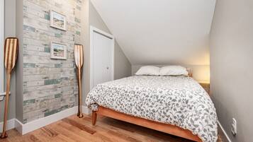 A bedroom with a sloped ceiling, a wooden floor, a bed with a patterned bedspread, a wall with a wooden plank design, and two wooden paddles leaning against the wall.