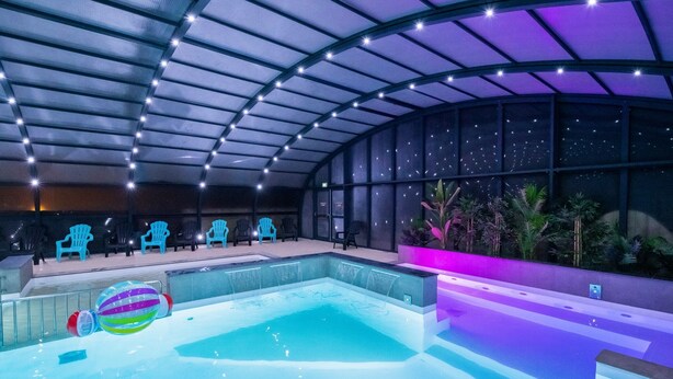 A modern indoor pool area with a clear water surface, a large glass enclosure, and blue chairs along the poolside.