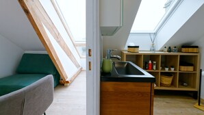 A loft-style room with a skylight, a green sofa, a kitchen area with a sink and wooden shelves.