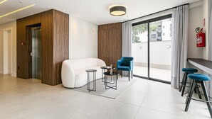 Een moderne hotellobby met een witte bank, een blauwe stoel, twee barstoelen, een kleine tafel en een brandblusser.