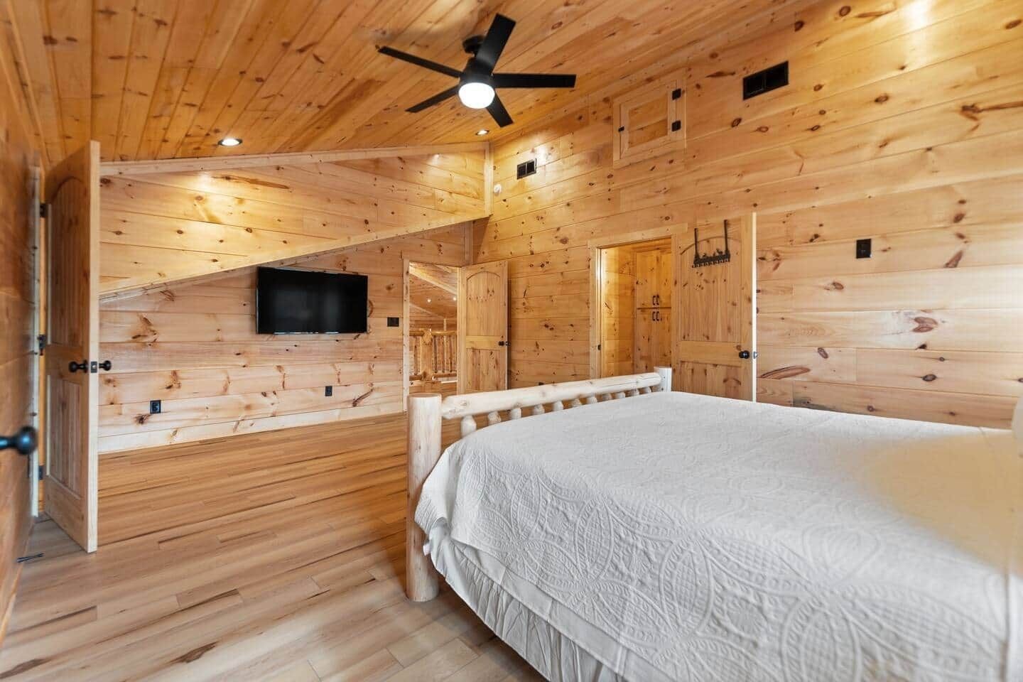 Une chambre à coucher dans une cabane en bois, avec un lit, un ventilateur de plafond, un téléviseur fixé au mur et une porte donnant sur une autre pièce.