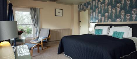 A bedroom with a bed, a chair, a desk, a lamp, a window with curtains, and a wall with a floral pattern.