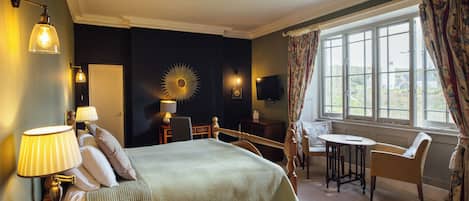 Habitación de hotel con cama, lámparas de noche, una mesita, una silla, televisor y ventana con cortinas.