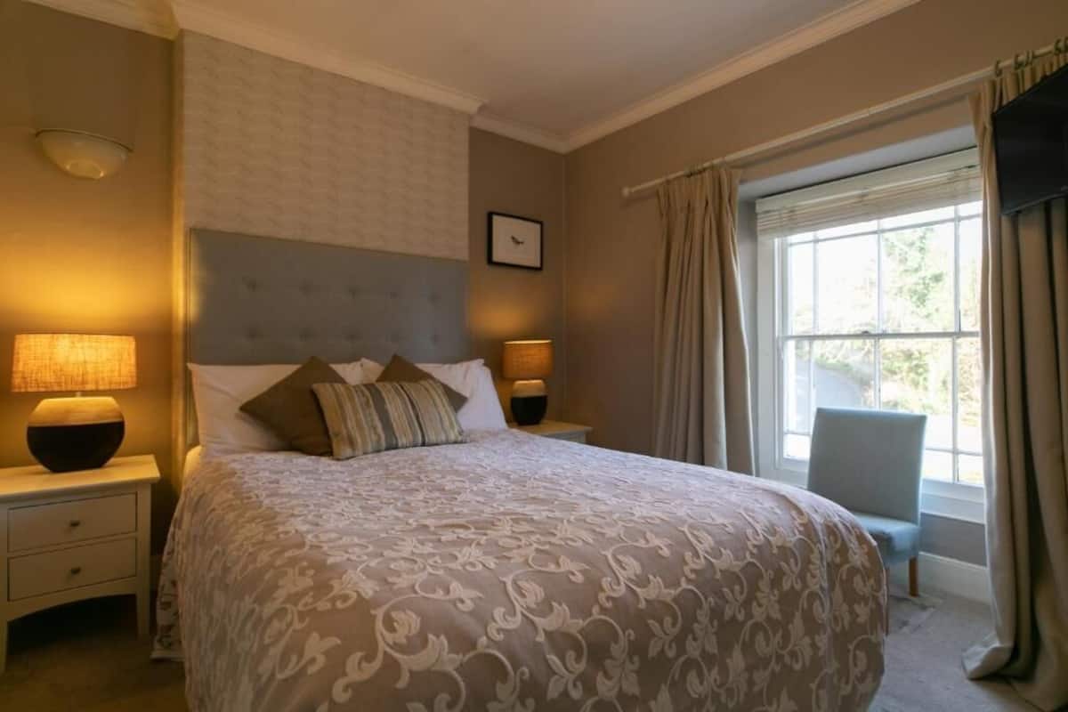 A bedroom with a large bed, two bedside tables, a chair, a window with curtains, and a wall-mounted picture.