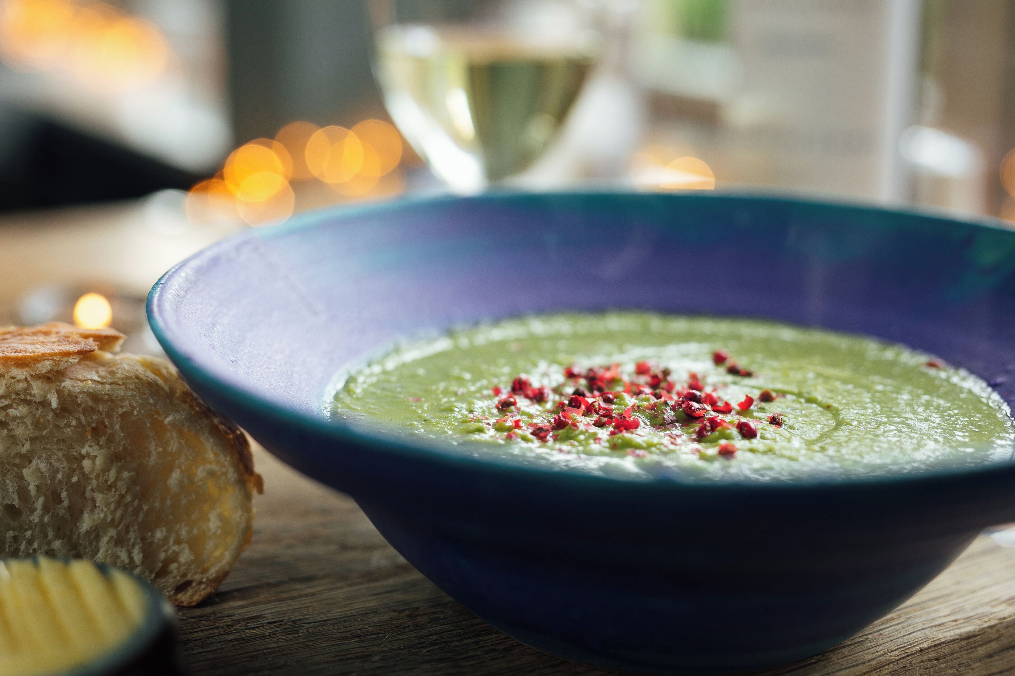 Un tazón de sopa verde adornado con hojuelas de chile rojo, sobre una mesa de madera con una vela al fondo.