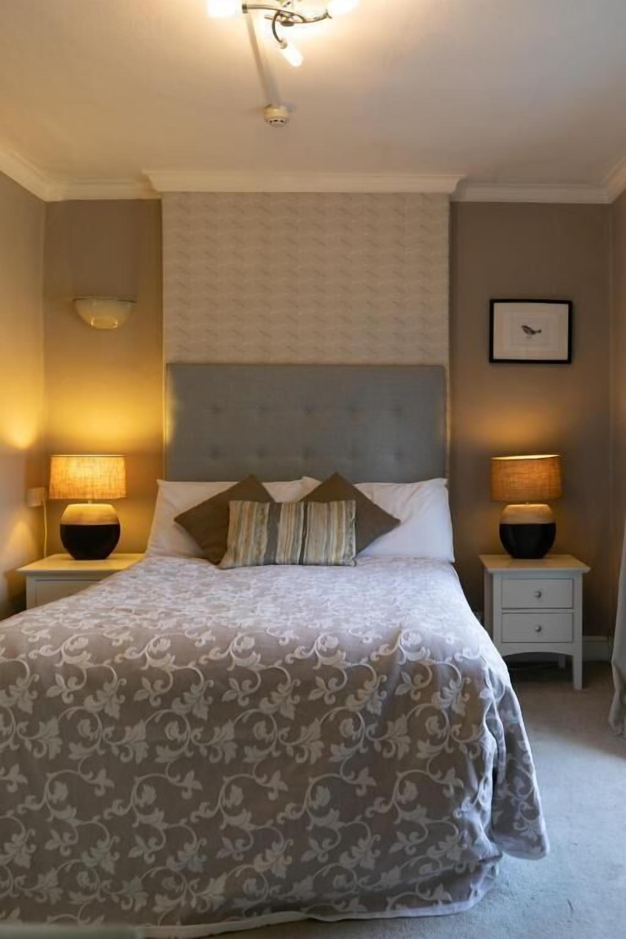 Ein ordentlich eingerichtetes Schlafzimmer mit einem Bett, zwei Nachttischen, einer Wandleuchte und einem gerahmten Bild.