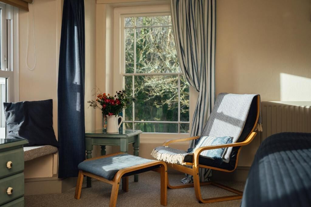 Una habitación con una cama, una silla, una mesita con un jarrón de flores y una ventana con cortinas.