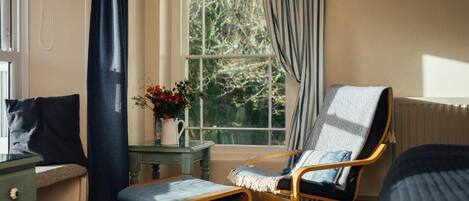 Una habitación con una cama, una silla, una mesita con un jarrón de flores y una ventana con cortinas.