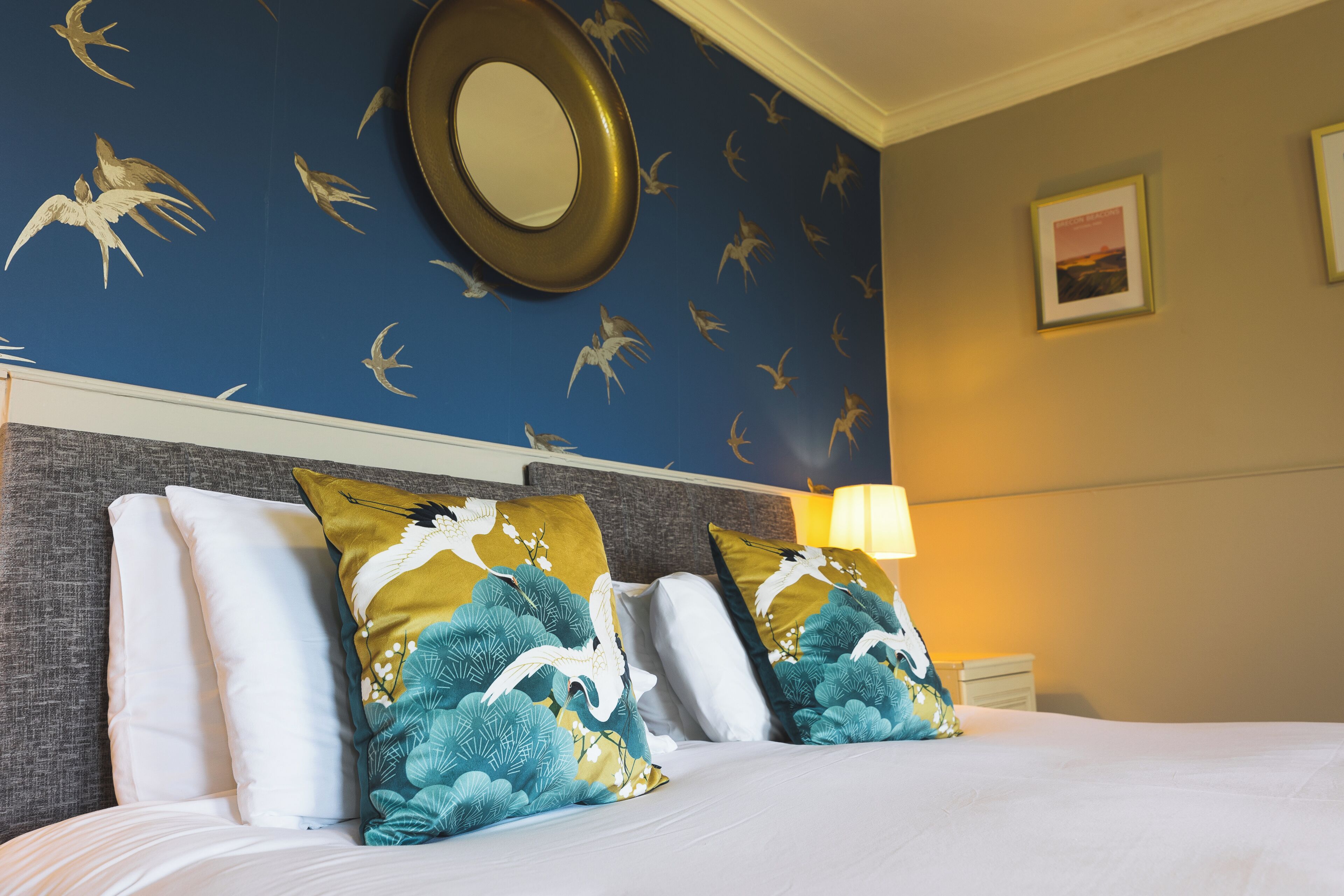Une chambre d’hôtel avec un lit, des oreillers, une tête de lit, un mur orné d’un motif d’oiseaux, un miroir rond et un tableau encadré.
