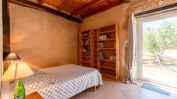 Een slaapkamer met een bed, een nachtkastje, een lamp, een boekenkast en een raam met uitzicht op de omgeving.