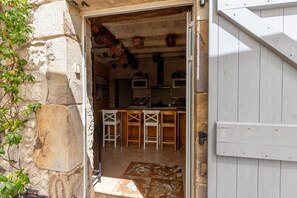 Une façade en pierre rustique, avec une porte en bois donnant sur une cuisine au sol carrelé et dotée de tabourets en bois.