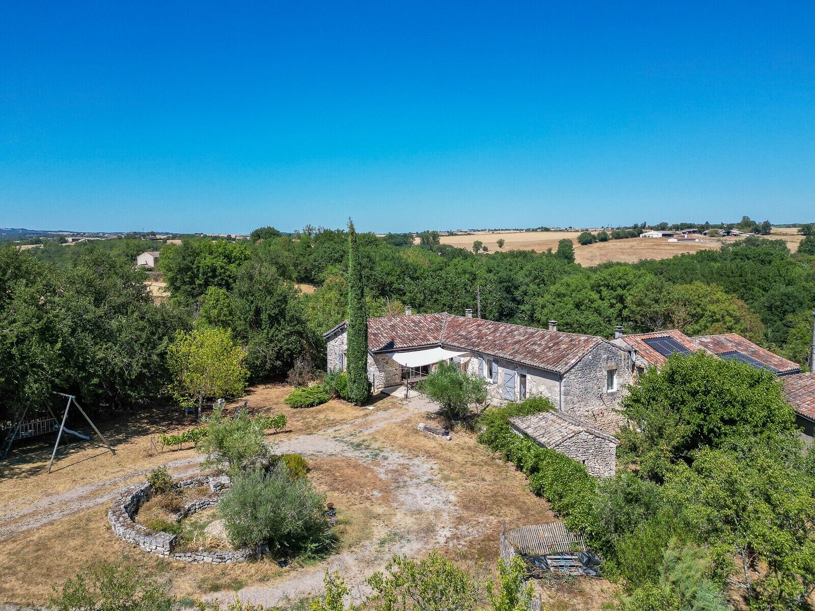 Une propriété rurale avec des bâtiments en pierre, un jardin circulaire également en pierre et une végétation luxuriante tout autour.