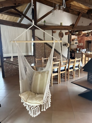 A room with a hanging chair, wooden dining table, chairs, and a kitchen area. - Gîte L'Arc-en-Ciel: Spacious Charming House with Central Fireplace (Bournazel)